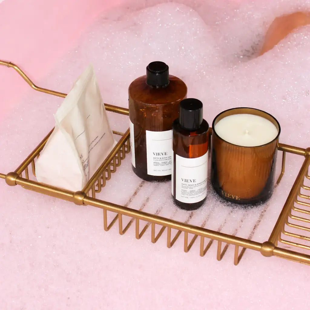 Porte-bains en métal doré contenant des bouteilles en verre ambré et une bougie avec des étiquettes blanches.